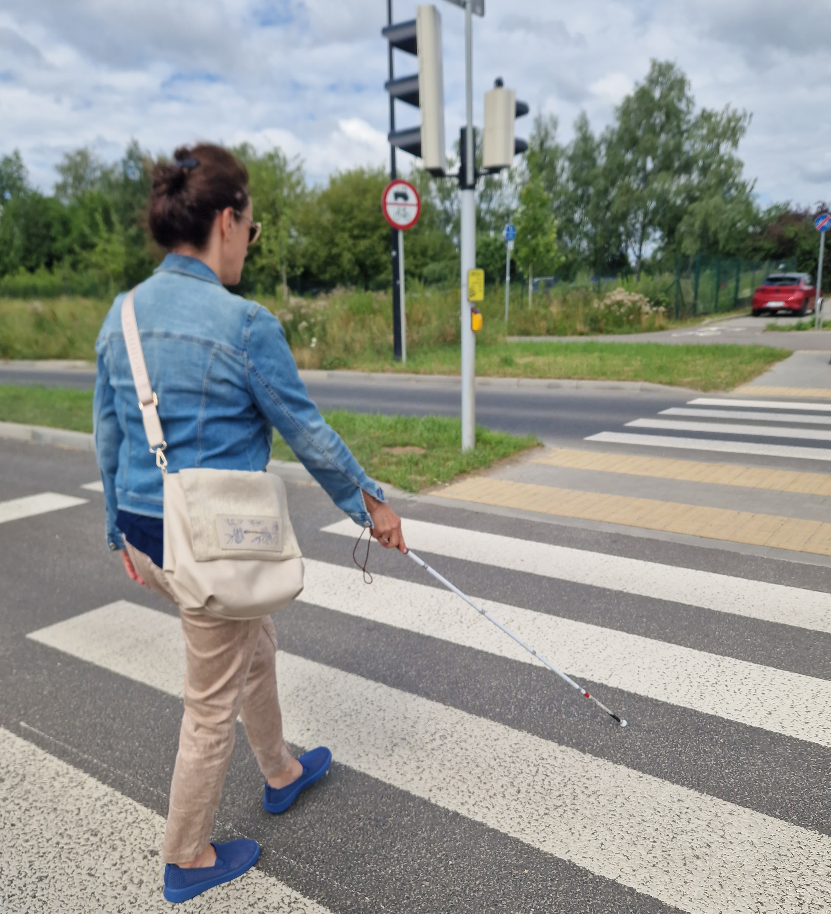 Kobieta ubrana w dżinsową kurtkę i jasne spodnie trzymająca laskę dla niewidomych przechodzi przez przejście dla pieszych
